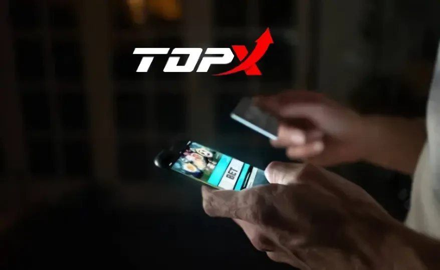 Duas mãos usando celulares no escuro, um deles com tela iluminada, e o logotipo da TopX ao fundo
